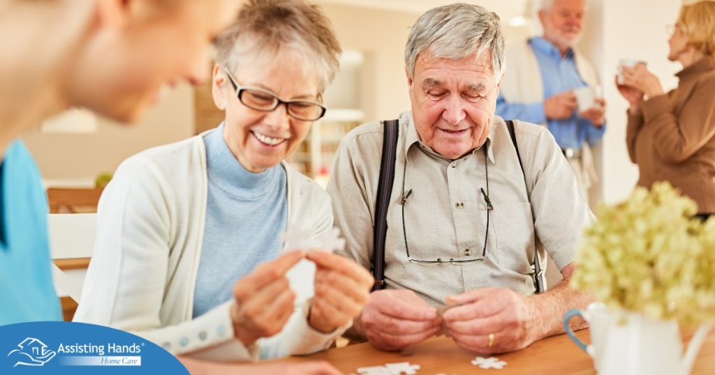 An older couple works on a puzzle with a caregiver, representing the kind of activity that helps those with dementia and also representing Alzheimer’s and Brain Awareness Month.