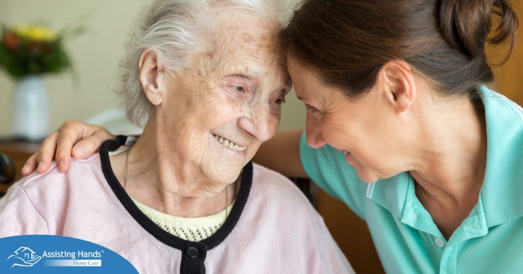 A caregiver lovingly embraces a senior woman while they both smile, representing the kind of warm disposition that can help when dealing with dementia-related repetition.