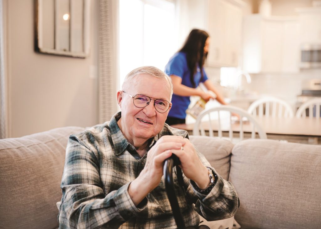 Senior man with a cane smiling while sitting on a couch as his caregiver provides home assistance in the background.