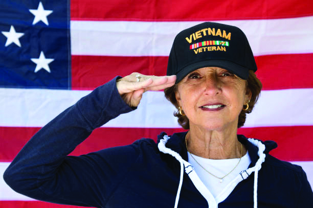 Woman Vietnam veteran in a navy jacket and “Vietnam Veteran” cap saluting in front of an American flag.