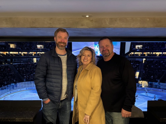 Tim and Lisa from Assisting Hands Home Care stand with Sean from Northern Utah Rehabilitation Hospital at a hockey game overlooking the ice.