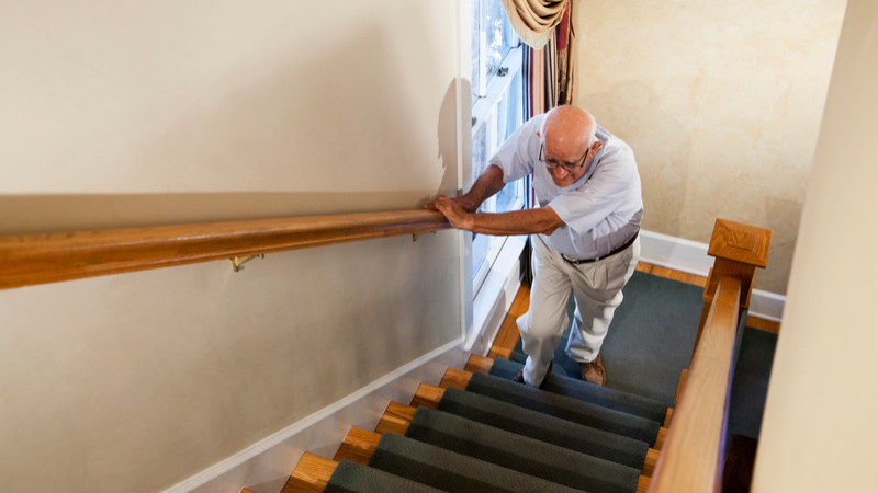 Older man holding a stair railing while walking up steps at home