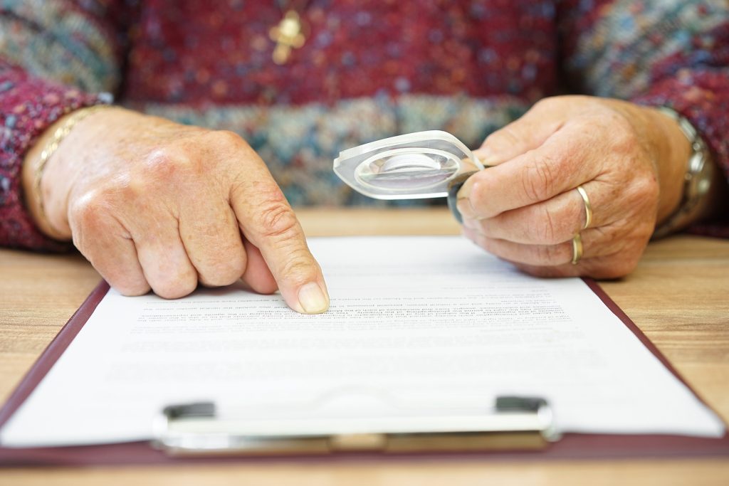An elderly person carefully reviewing documents with a magnifying glass, pointing to the fine print of what appears to be an insurance policy. Understanding your long-term care insurance in Murray, UT is the first step toward making the most of your benefits. A trusted provider of senior home care in Murray, UT can help guide you through the process.