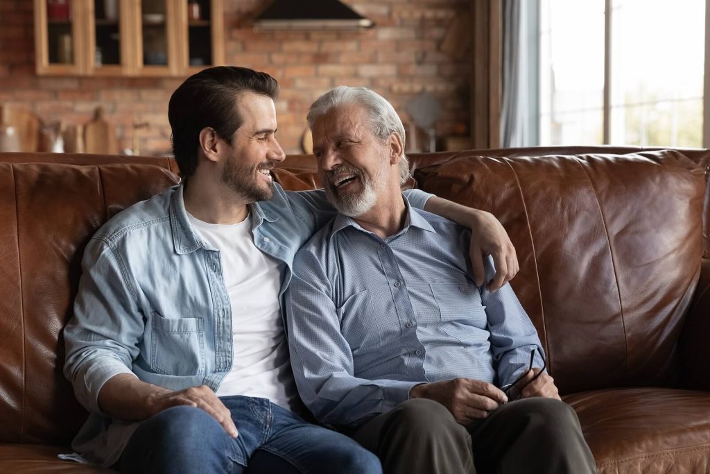 An adult son and his elderly father share a warm, joyful moment together on the couch, reflecting the meaningful connection that family caregiving is built on. Respite care services in Murray, UT help preserve these relationships by giving family caregivers the rest they need to show up fully for their loved ones. When caregivers are supported, moments like these become more possible.