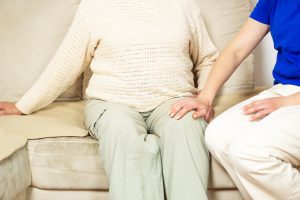 A caregiver places a reassuring hand on the knee of an elderly person seated on a couch, representing the steady, in-home presence that helps prevent caregiver burnout in Murray, UT. Professional Alzheimer's care in Murray, UT gives family members the relief and support they need, while ensuring their loved one receives consistent, dignified care at home.