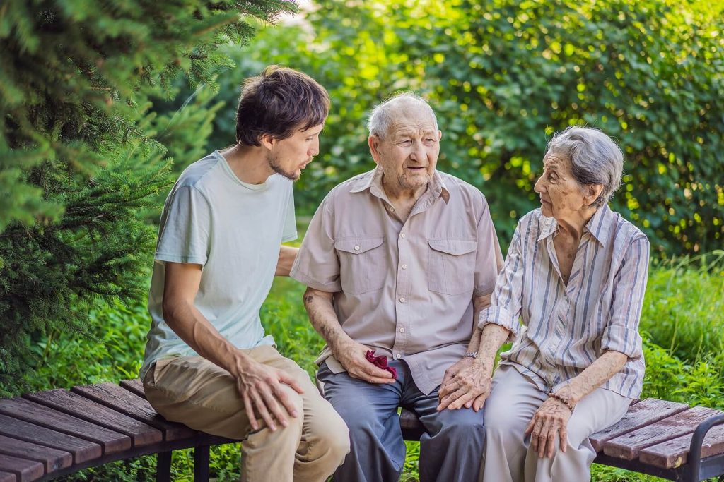 A young man sits with his elderly grandparents on an outdoor bench, engaged in a warm and caring conversation — the kind of moment that motivates families to seek Alzheimer's care in Murray, UT before memory loss progresses. Having the support of a trusted senior caregiver in Murray, UT can help families like this one navigate the journey together without facing it alone.