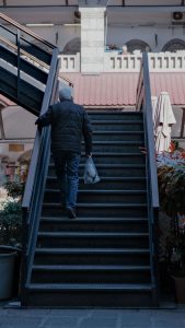 An older man carefully climbs a staircase while carrying groceries, a common everyday risk that highlights the importance of fall risk home care in Utah, fall risk home care Utah County, and fall prevention for seniors in Murray, UT. 