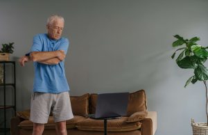 An older man stretches his arm while following an exercise class on his laptop at home, a routine supported by in-home care for seniors in Murray, UT, fall risk home care Utah County, and fall prevention for seniors in Murray, UT. 