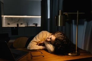 A woman has fallen asleep at her desk late at night beside her laptop and glasses, worn down after another long day of juggling work and caregiving. It's a familiar picture of burnout in caregivers Murray, UT, and a reminder that respite care near me and respite care Murray can help her rest. 
