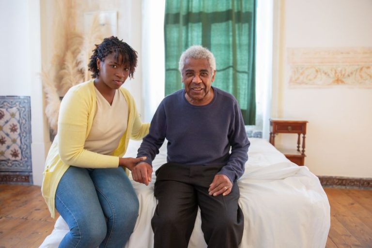 Caregiver helping an older man sit up safely on the edge of the bed