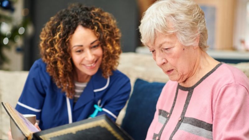 Assisting Hands Home Caregiver With Dementia Senior Woman at home reding book Assisting Hands Home Caregiver With Dementia Senior Woman at home reding book