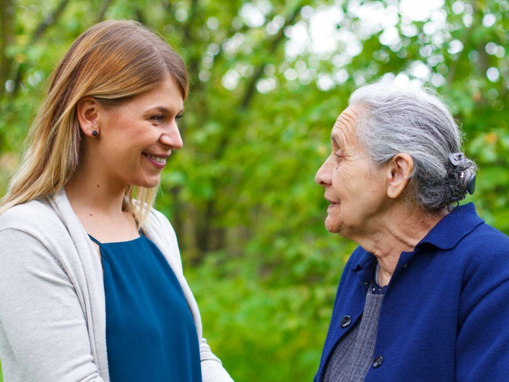 A caregiver smiles at an elderly woman as they walk through a forest. Coordination of Care in Wellesley
