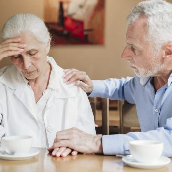 A husband and wife sitting at a kitchen table. Wife has hand on forehead and husband reassures wife with hand on shoulder. Alzheimer’s and Dementia Care in Waltham