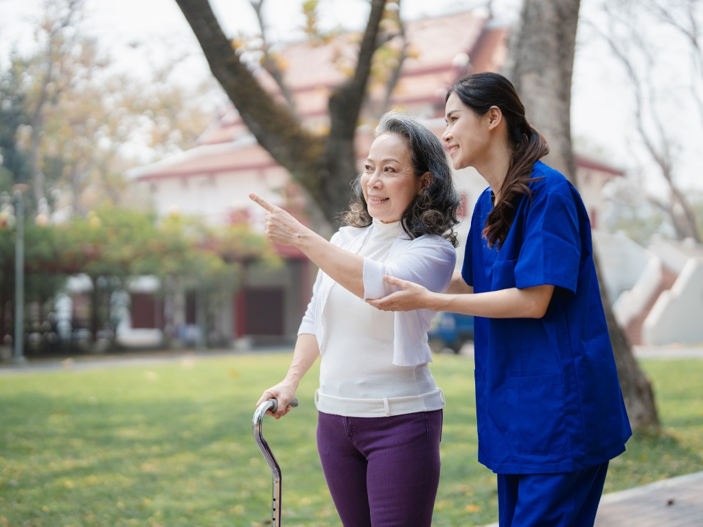 A senior woman using a cane enjoys a walk in the park with a supportive caregiver in blue scrubs, illustrating quality Personal Care & Companionship in Billerica