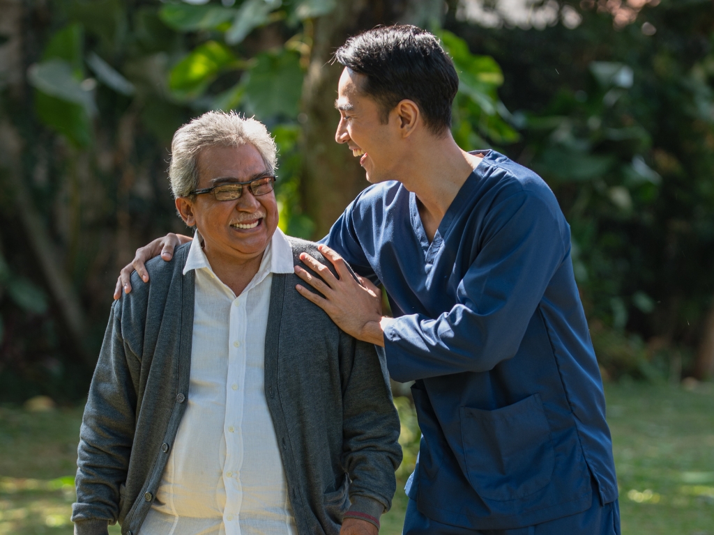 A caregiver in blue scrubs supports and smiles with an elderly man outdoors, symbolizing compassionate Coordination of Care in Billerica.