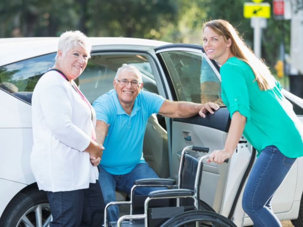 Transportation-Services-in-Bedford A caregiver assists an elderly man into a car while another caregiver prepares his wheelchair, representing safe and reliable transportation care services in Billerica.