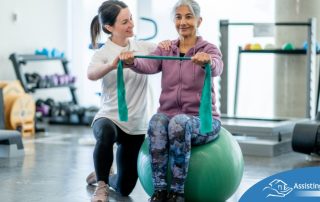 A care provider helps an older woman exercise with a resistance band and an exercise ball, representing how exercise can help with senior fall prevention.