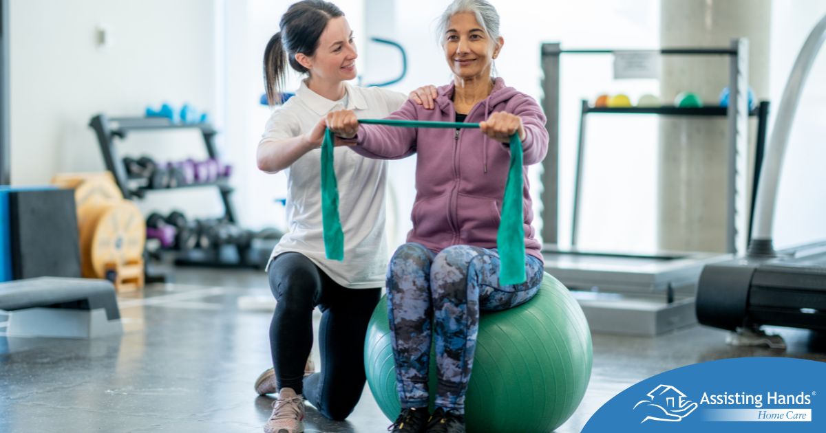 A care provider helps an older woman exercise with a resistance band and an exercise ball, representing how exercise can help with senior fall prevention.