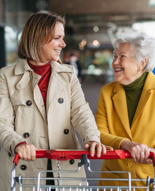 Grocery Shopping and Errands Services in Newton, MA (1) Smiling caregiver and senior client pushing a shopping cart together, Grocery Shopping and Errands Services in Newton, MA