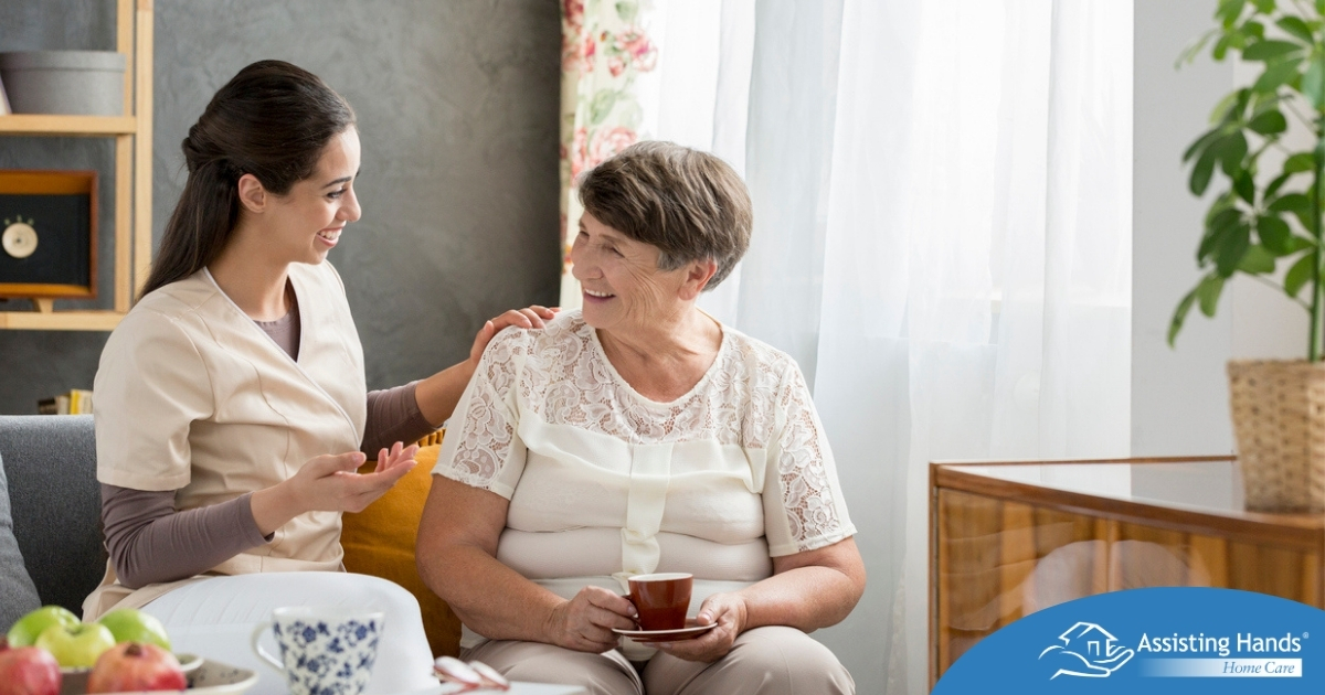 Home Care vs Assisted Living Where Caregiving Work Fits in Today’s Families Smiling caregiver with her hand on an older woman’s shoulder as they share tea on the couch, showing home care vs assisted living
