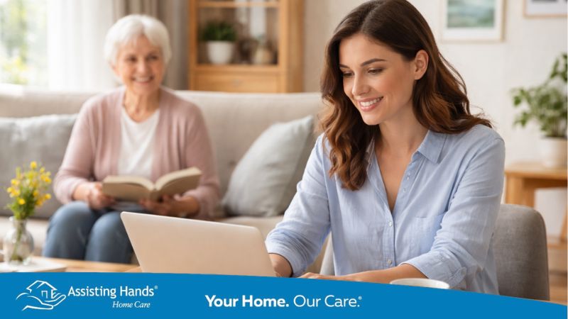 A middle-aged adult daughter works on a laptop at a wooden table, smiling, while her elderly mother sits nearby on the sofa reading a book.