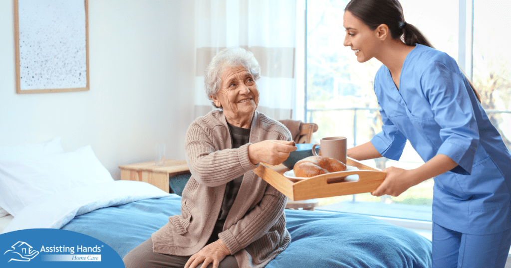 A professional caregiver smiles and provides food to an older adult in her care, representing the compassionate care needed for those with dementia.